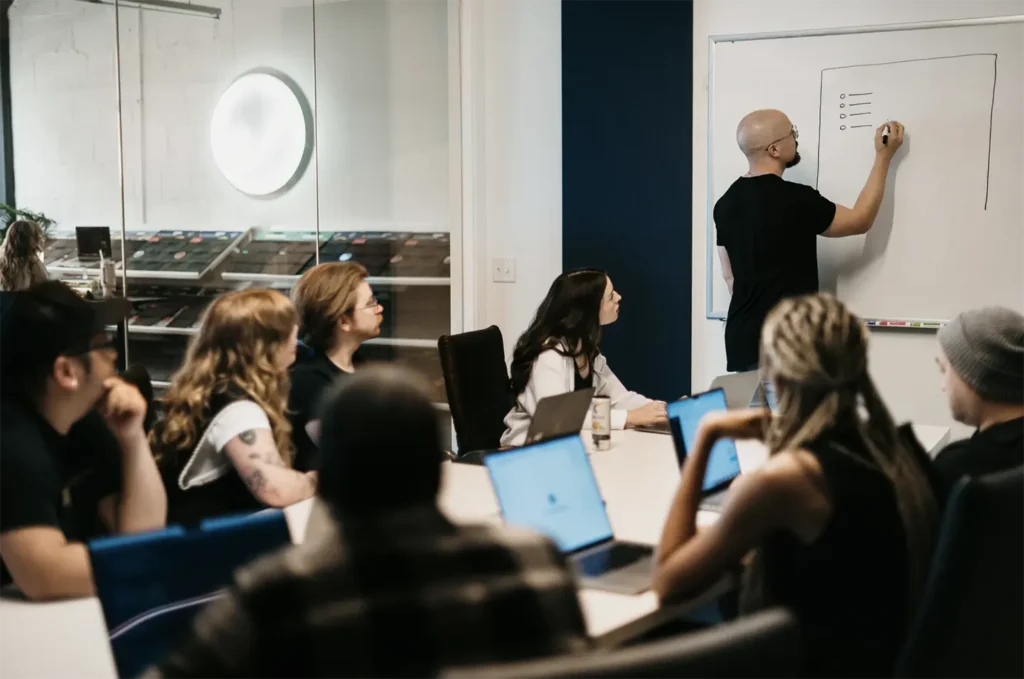 A woman with long dark hair, wearing a white shirt and layered necklaces, sits at a desk engaged in a phone call. She smiles as she works, with a laptop open before her and a mug and mouse nearby, embodying her daily routine at Hook Agency.