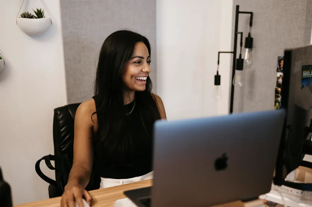 A woman with long dark hair, wearing a white shirt and layered necklaces, sits at a desk engaged in a phone call. She smiles as she works, with a laptop open before her and a mug and mouse nearby, embodying her daily routine at Hook Agency.