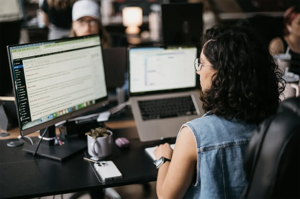 A woman with long dark hair, wearing a white shirt and layered necklaces, sits at a desk engaged in a phone call. She smiles as she works, with a laptop open before her and a mug and mouse nearby, embodying her daily routine at Hook Agency.