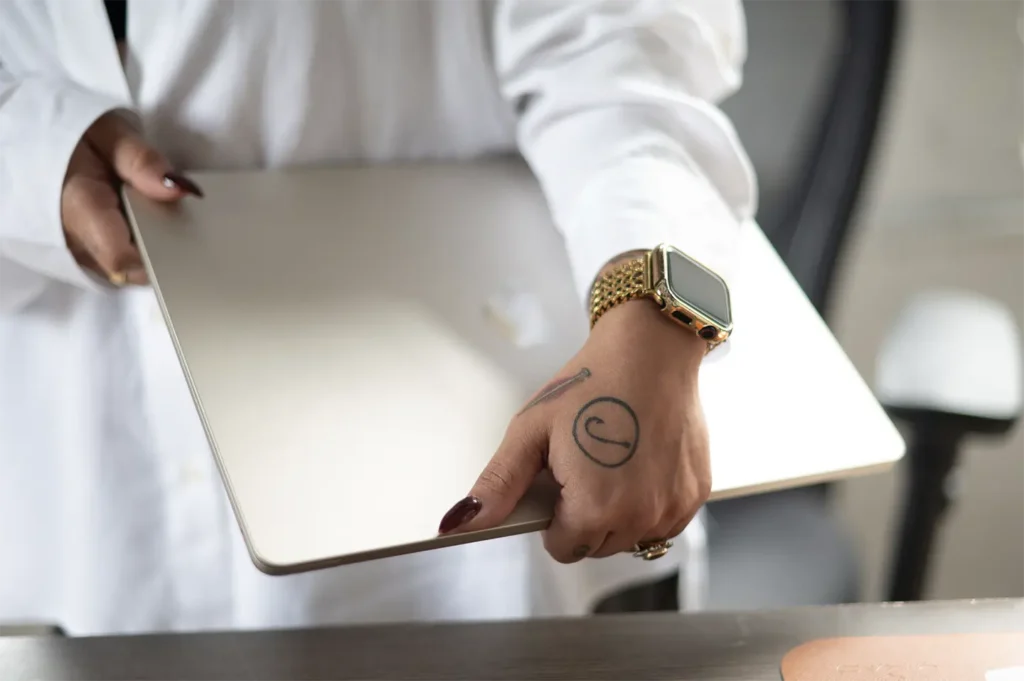 A woman with long dark hair, wearing a white shirt and layered necklaces, sits at a desk engaged in a phone call. She smiles as she works, with a laptop open before her and a mug and mouse nearby, embodying her daily routine at Hook Agency.